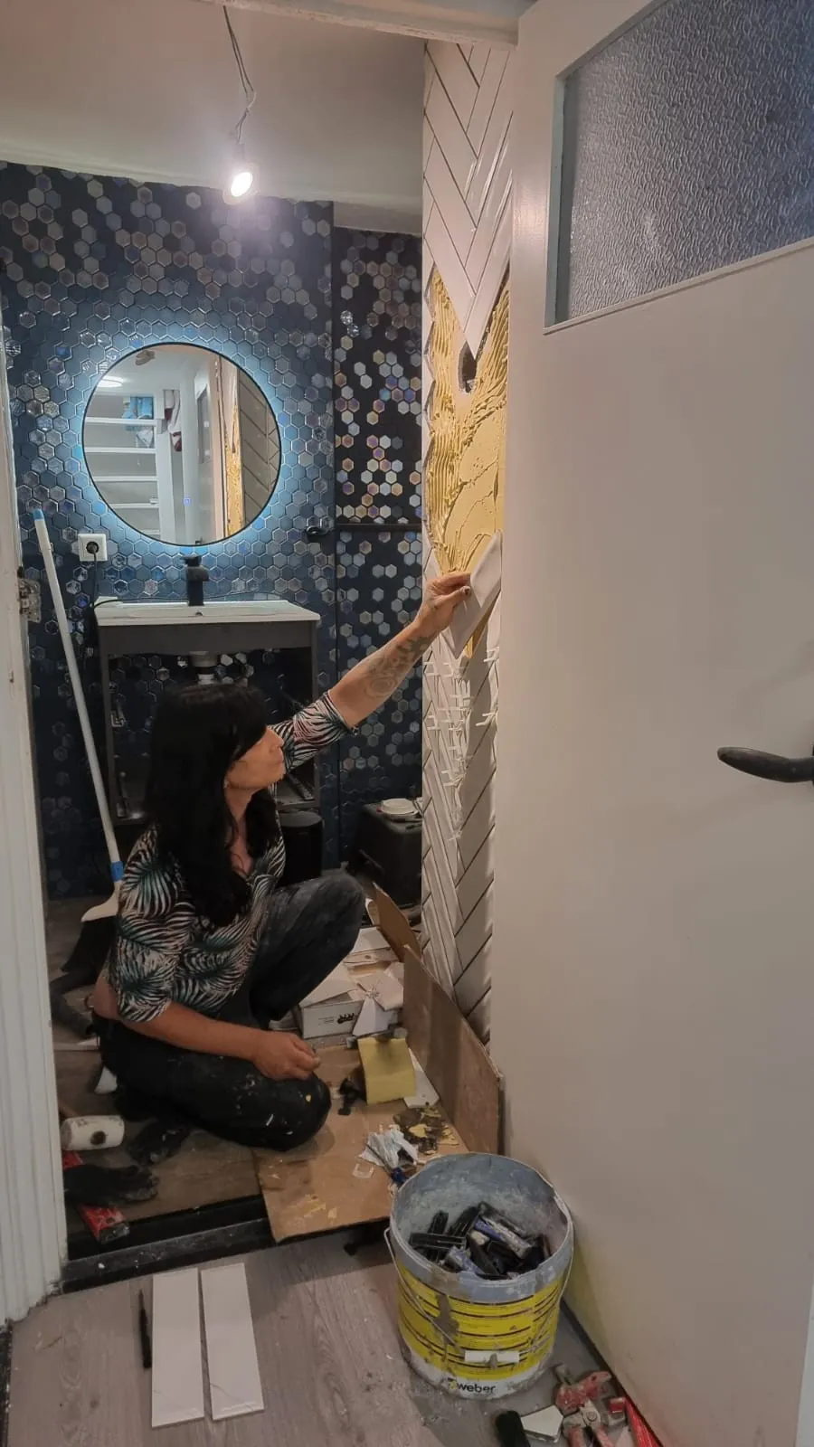 Badkamerrenovatie in uitvoering in Sittard: Diana zet wandtegels in visgraatpatroon, met blauw hexagon-mozaïek en verlichte spiegel.