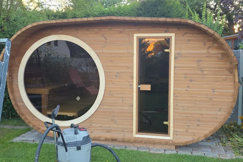 Buitensauna in hout met rond patrijspoortraam en verlichte deur, in een tuin.