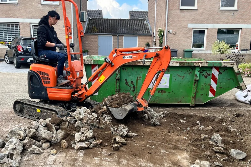 Minigraafmachine breekt de oude bestrating op, puin gaat rechtstreeks de container in.